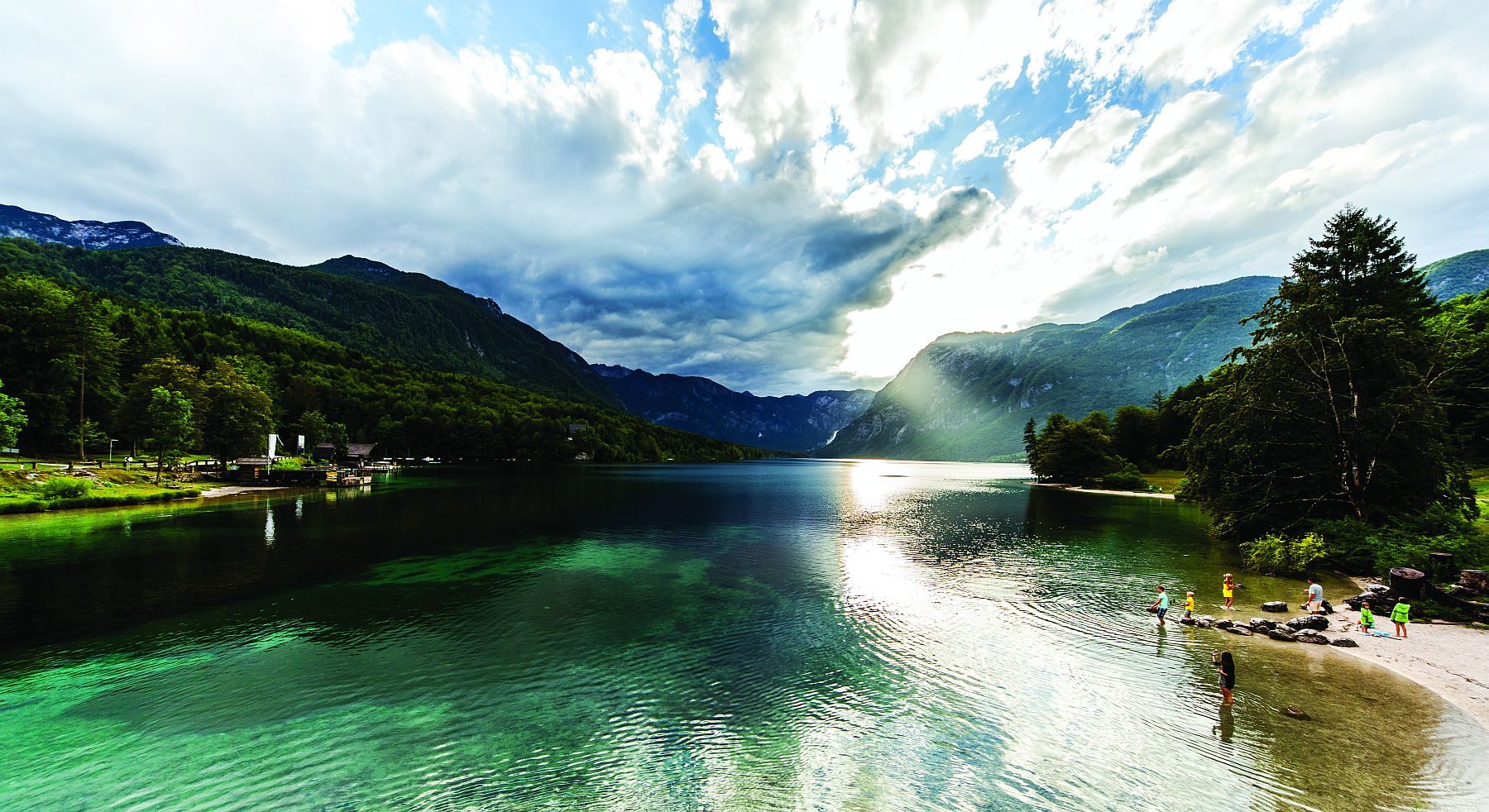 Kirkkaana kimmeltävä Bohinj-järvi. Lapsia kahlaamassa rantavedessä.
