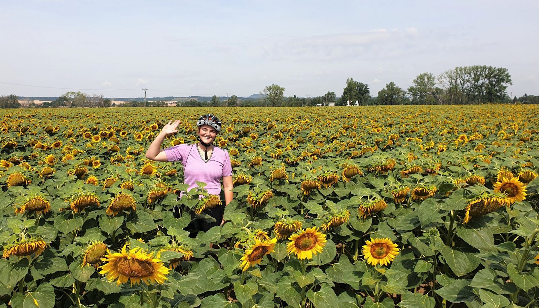Nainen vilkuttaa iloisesti auringonkukkapellon keskeltä.