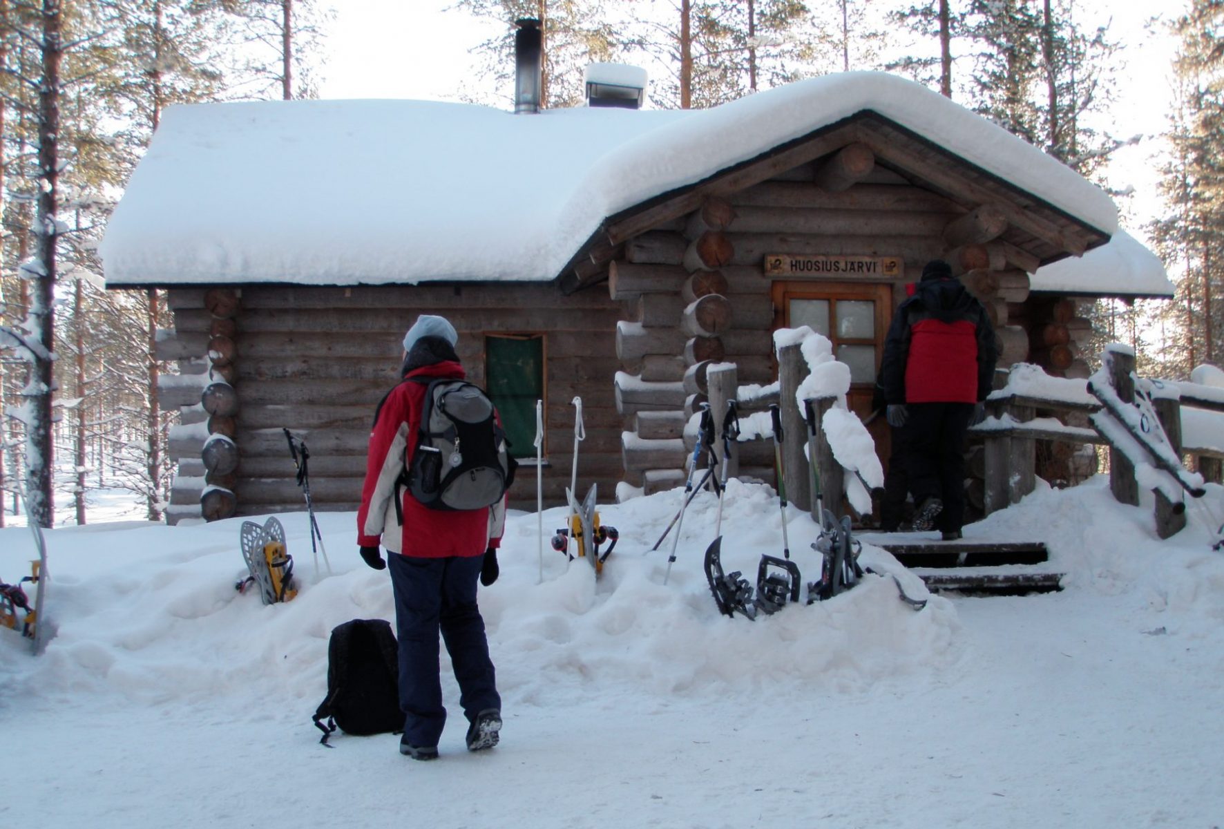Lumisen maiseman ympäröimä eräkämppä, jonka pihalla kaksi miestä.