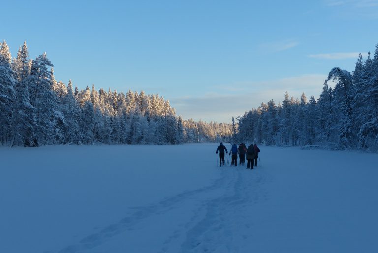 Ryhmä lumikenkäilijöitä tammikuun "sinisessä hetkessä" järven jäällä. Ympärillään kauniin kuuraiset puut.
