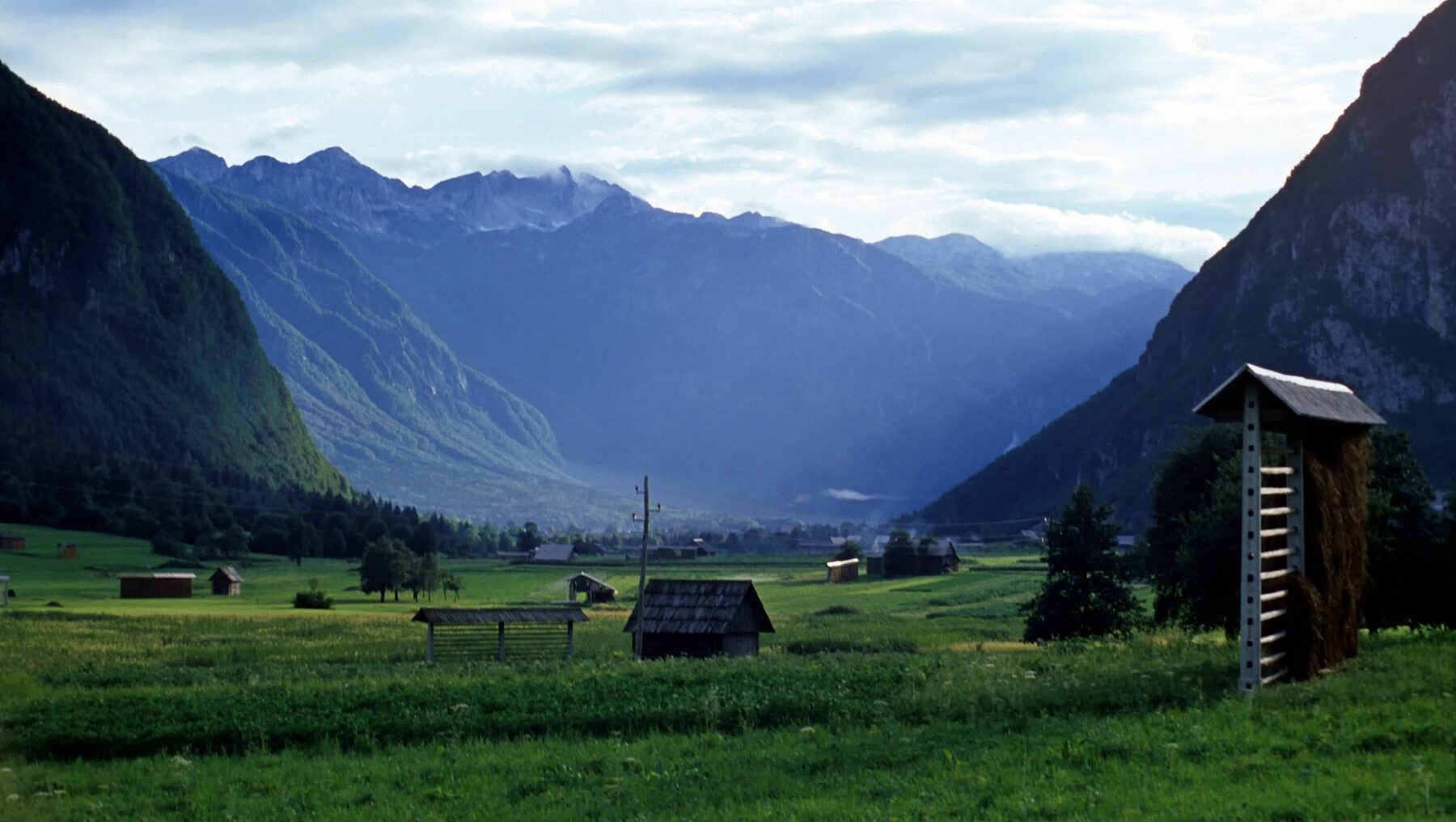 Vuorten ympäröimä niitty heinälatoineen aamunkoitteessa.