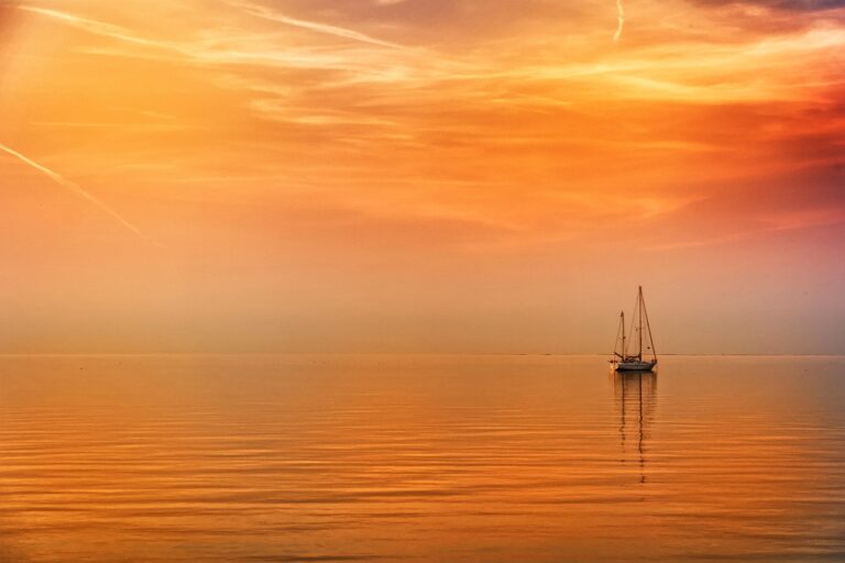 Pieni purjevene lipuu tyynellä järvellä ilta-auringon oranssiksi värjäämässä maisemassa.