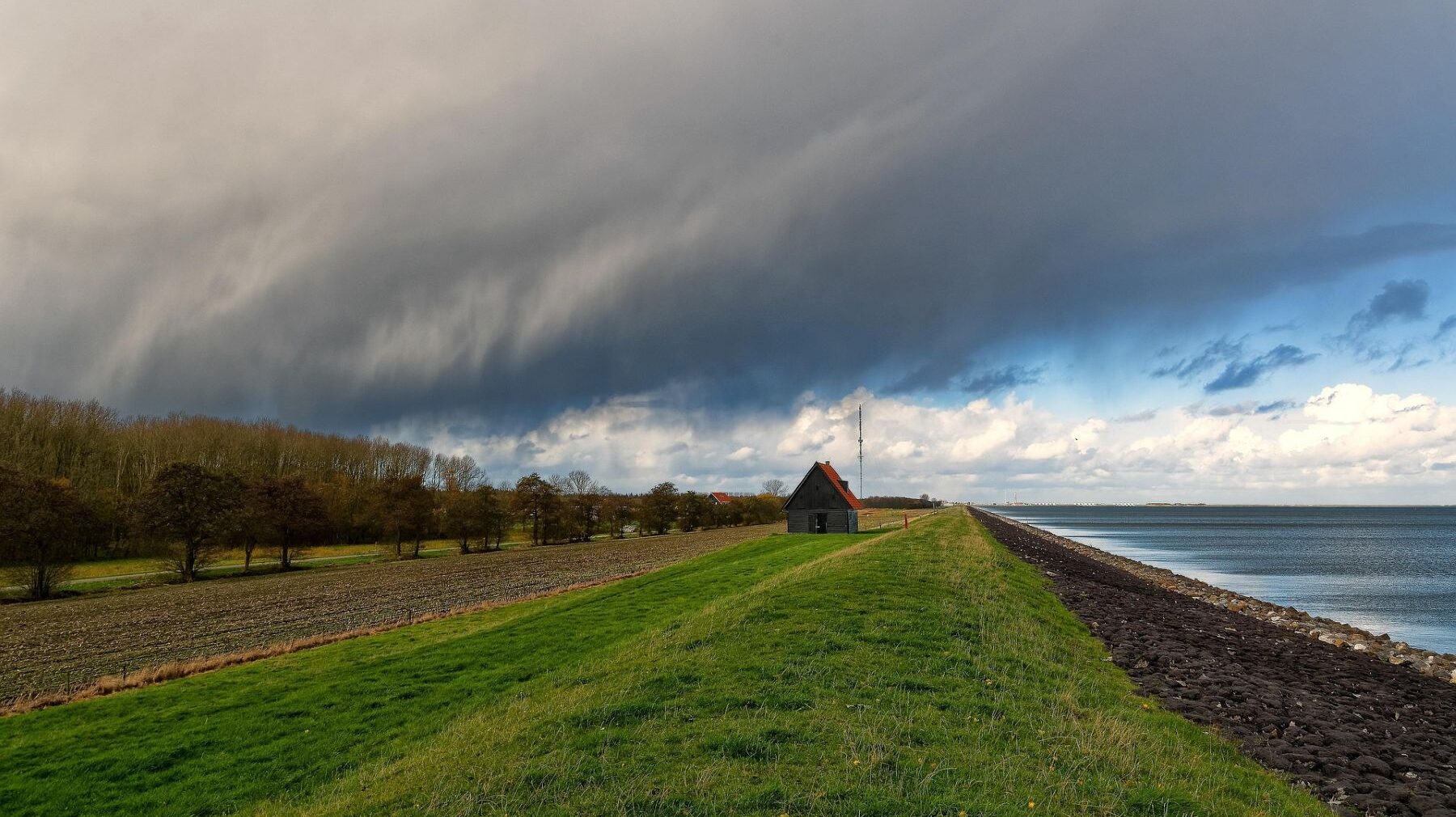 Rantavallin päällä sijaitseva pieni mökki tummien pilvien alla. Vasemmalla puita ja peltoa, oikealla rauhallinen merimaisema