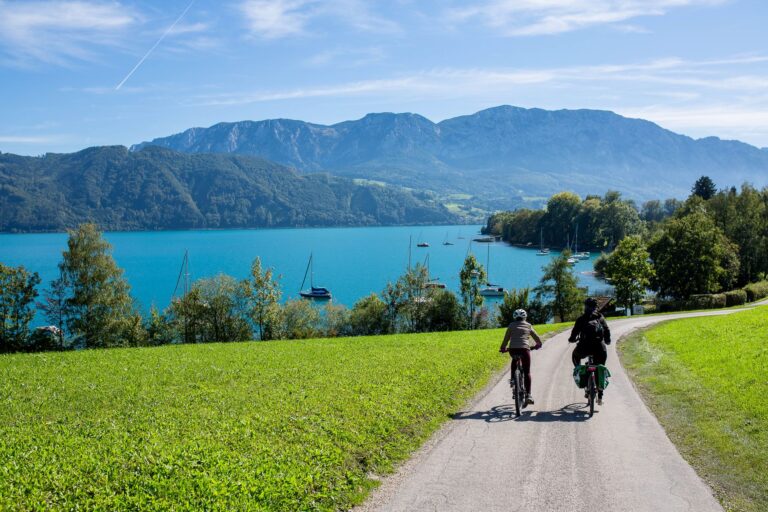 Kaksi pyöräilijää ajaa vihreän niityn reunustamaa pyörätietä kohti järveä.